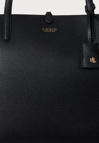 Μαύρη δερμάτινη τσάντα tote με υφή, δύο λαβές, χρυσό λογότυπο εμπρός και αντίστοιχη ετικέτα. Λείος σχεδιασμός με κομψό φινίρισμα.