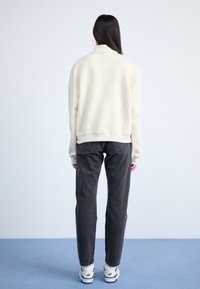 Felpa beige chiaro con collo alto e polsini a coste, abbinata a jeans grigio scuro e scarpe da ginnastica bianche, vista posteriore.