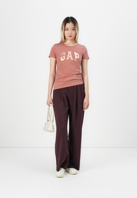 Pink t-shirt with "GAP" logo in light beige, paired with flowy dark maroon pants and white sneakers. Model holds a small white handbag.