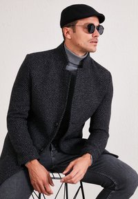 Cappotto nero con texture, colletto alto, silhouette aderente e due bottoni. Indossato sopra un dolcevita grigio; abbinato a jeans e occhiali da sole.