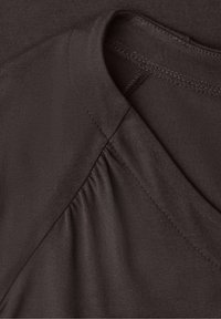 Gros plan sur un tissu marron foncé montrant l'encolure du vêtement et les détails des coutures avec de légers fronces près de l'épaule.