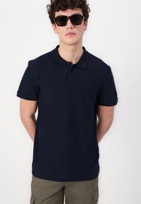 Tamno plava polo majica s teksturiranom tkaninom, kratkim rukavima, placket s tri gumba i standardnim krojem. Uključuje zelenne cargo kratke hlače i sunčane naočale.