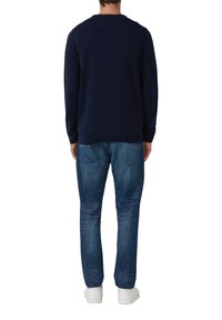 Uomo in maglione blu navy, jeans blu con tasche posteriori e sneakers bianche, in piedi di spalle su uno sfondo bianco.