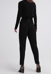 Mono negro con un diseño de top corto, textura suave y cierre de cremallera con herrajes dorados. Combinado con sandalias negras de tacón grueso.