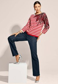 Rood-wit gestreepte trui met een zwarte bloemenprint op één mouw, gecombineerd met donkere jeans en zilveren hoge hakken. Model poseert op een kubus.