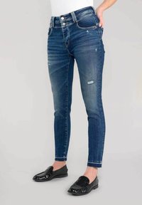 Dunne, donkerblauwe denim jeans met lichte slijtage, een hoge tailleband en vijf zakken; gecombineerd met zwarte instappers.