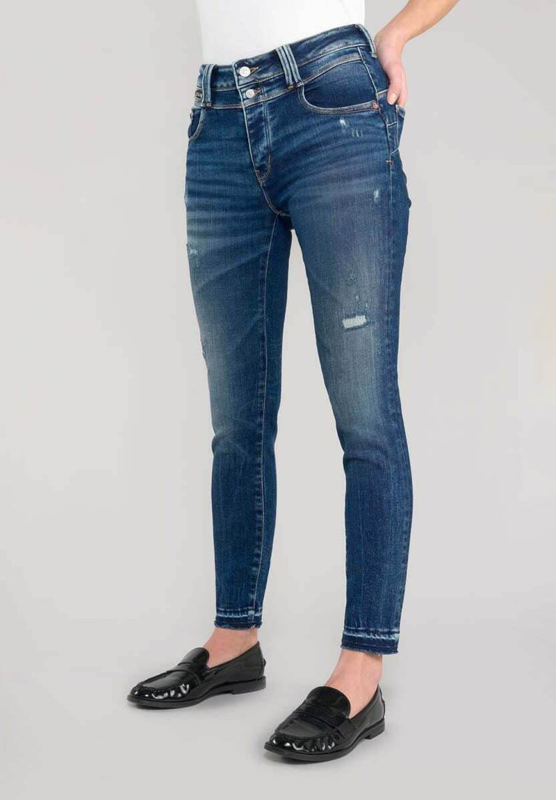 Dunne, donkerblauwe denim jeans met lichte slijtage, een hoge tailleband en vijf zakken; gecombineerd met zwarte instappers.