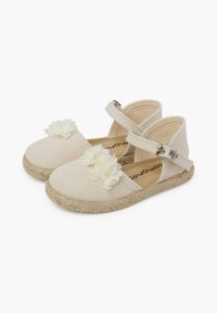 Béžové plátěné espadrilky s bílými květinovými akcenty, popruhem kolem kotníku a tkanou jutovou podrážkou. Zaoblený design špičky s texturovaným povrchovým materiálem.