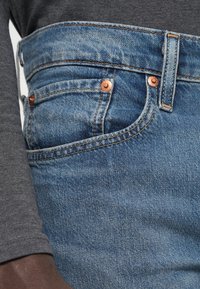 Traperice u izblijedjeloj plavoj nijansi, s mjedenim detaljima, ravnog kroja i bočnom džepom. Tekstuirana tkanina s vidljivim šavovima.