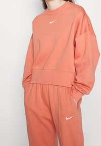 Korallfärgad sweatshirt med hög krage, drop shoulders och vit Nike-logga. Matcherande avslappnade sweatpants. Mjuk tygtextur.