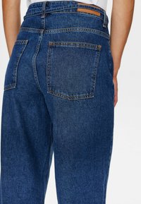 Tummansiniset farkut, joissa on rento istuvuus. Takana on kaksi taskua ja vyörengas on koristeltu ruskealla nahkakomponentilla. Klassiset tikkaukset näkyvät.