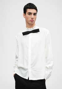 Bílá košile na knoflíky s volánkovým límcem a černou motýlkem. Košile má dlouhé rukávy a volný střih. Texturovaná bavlněná tkanina.