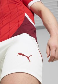 Sarkans sporta krekls ar smalkām līnijām, apvienots ar baltām šortiem, uz kuriem ir bordo Puma logo. Materiāls izskatās viegls un elpojošs.