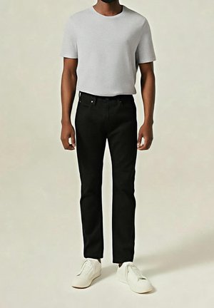 Mann trägt ein schlichtes hellgraues kurzärmeliges T-Shirt, schwarze Hose und weiße Turnschuhe und steht vor einem neutralen Hintergrund.