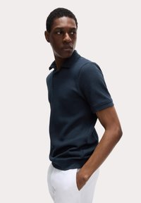 Camisa polo de un oscuro color marino con textura acanalada, mangas cortas y un cuello clásico. Combinada con pantalones blancos, enfatizando un look casual pero pulido.
