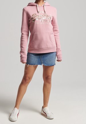 Vrouw draagt een roze met pailletten versierde hoodie met logo, een blauwe denim minirok en witte sneakers, staand tegen een effen achtergrond.