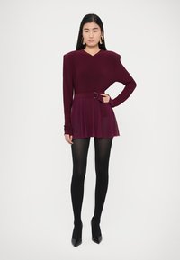 Vino crvena haljina dugih rukava s pripijenim korzetom, širokom suknjom, pojasevom u struku i suptilnim ramenskim jastučićima; uparena s crnim čarapama i štiklama.