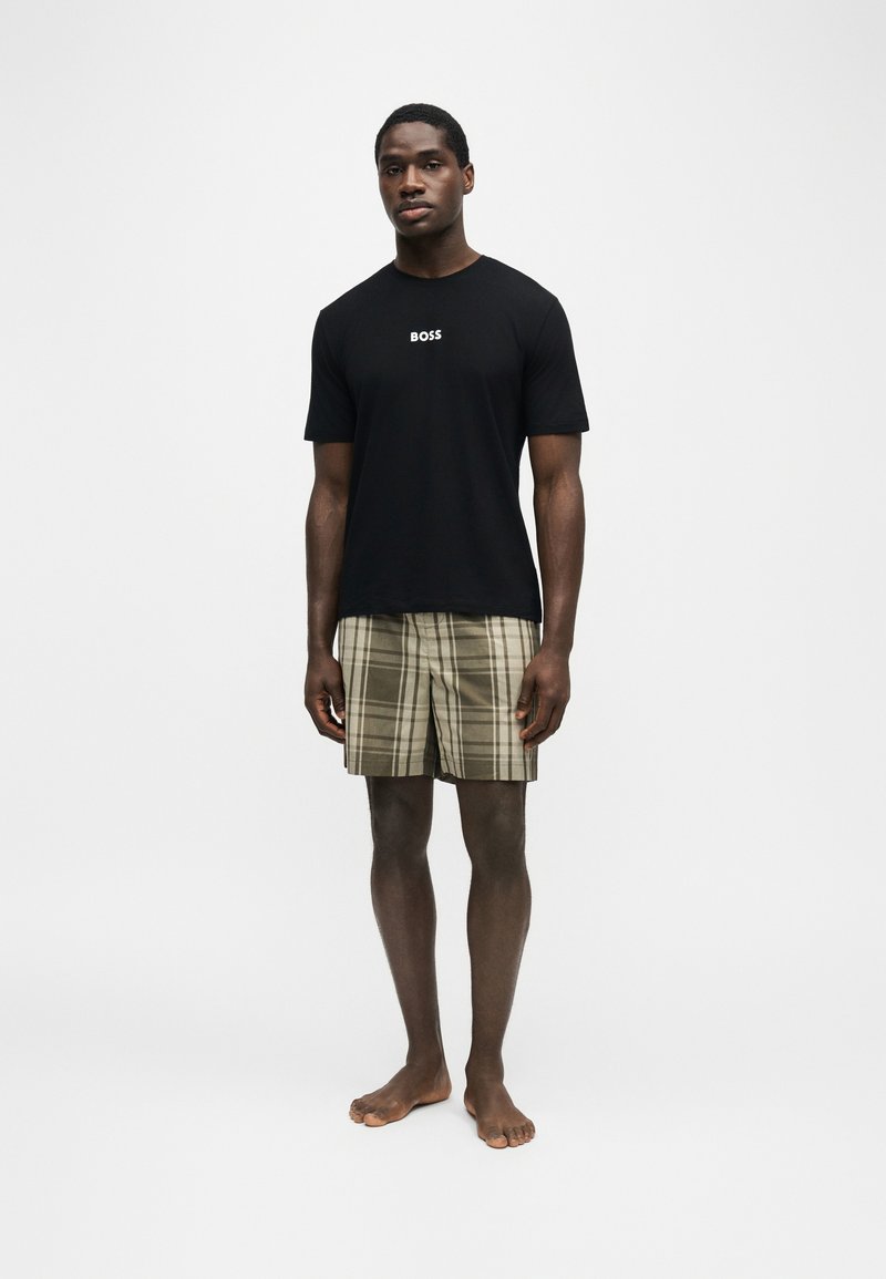 Homme debout pieds nus portant un t-shirt noir "BOSS" et un short à carreaux beige sur un fond clair uni.
