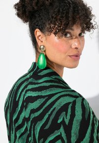 Abbigliamento a motivi zebra neri e verdi con una finitura testurizzata; grandi orecchini a goccia dorati e verdi esaltano l'aspetto generale.