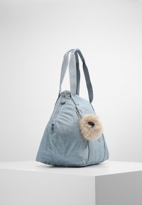 Sac fourre-tout en tissu bleu clair avec fermeture éclair, surface texturée, forme triangulaire et accessoire pompon duveteux amovible.