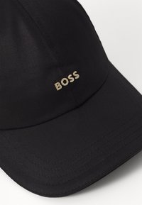 Căciulă neagră din țesătură cu o suprafață texturată, având un bor discret și curbat și un logo auriu "BOSS" pe partea din față.