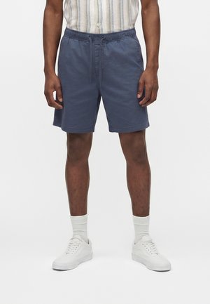 Mężczyzna w granatowych szortach ze ściągaczem, białych sportowych butach i białych skarpetkach, stojący na jednolitym białym tle.