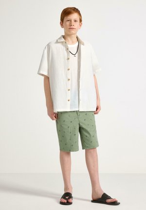 Dreng står iført hvid kortærmet skjorte med knapper, hvid t-shirt, grønne shorts med små palmetræsprint, sorte sandaler og et sort halskæde.