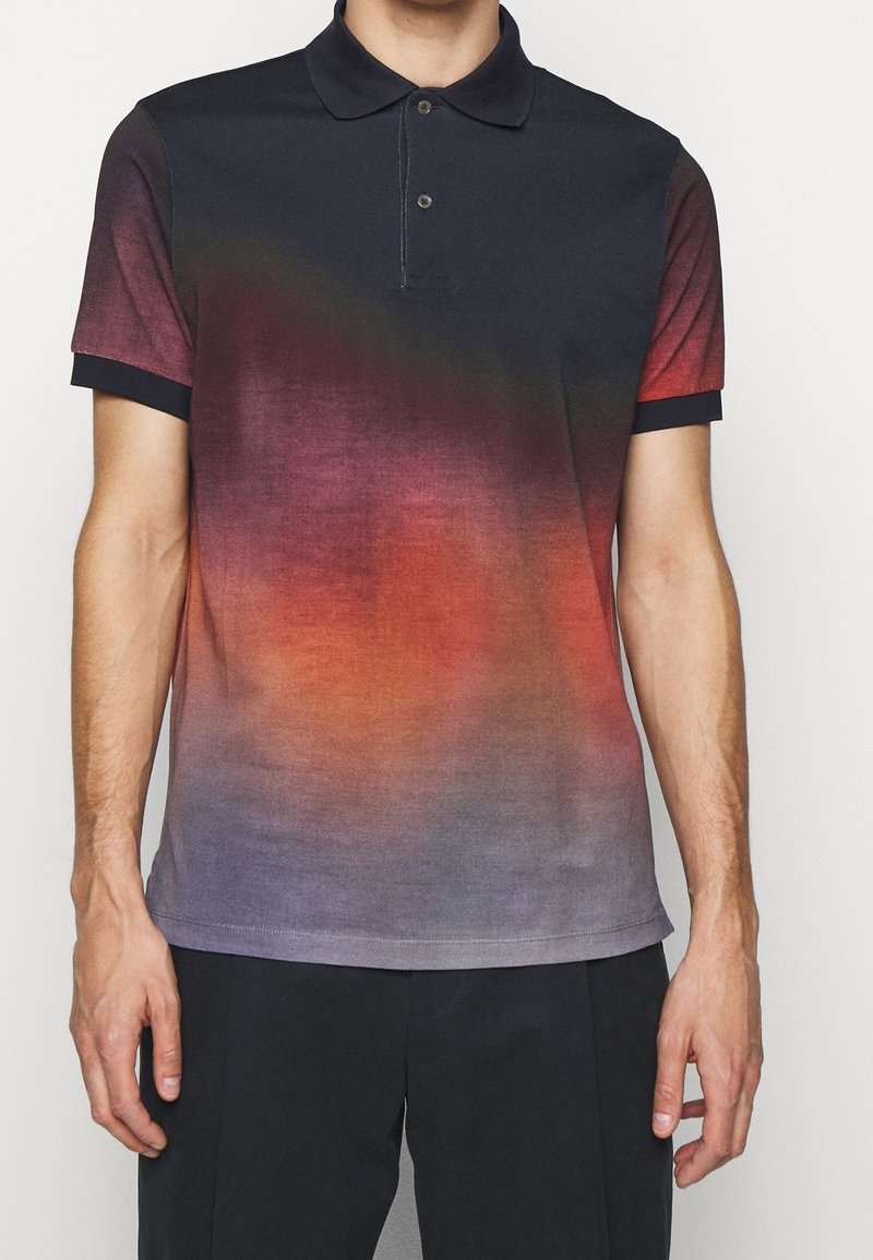 Homme portant un polo à manches courtes avec un motif dégradé allant du bleu foncé en haut au rouge et violet en bas, assorti à un pantalon noir.
