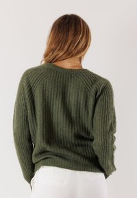 Donna con capelli castano chiaro che indossa un maglione texturizzato verde oliva e pantaloni bianchi, vista di spalle su uno sfondo semplice.