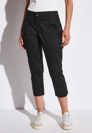 Mujer con pantalones negros cortos, zapatillas blancas y una camiseta blanca, posando frente a un fondo blanco liso.