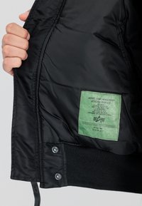 Sort nylonjakke med quiltet foer, der har et grønt vaskeanvisningsmærke, ribbede manchetter og trykknaplukninger. Design med fastgjort hætte.