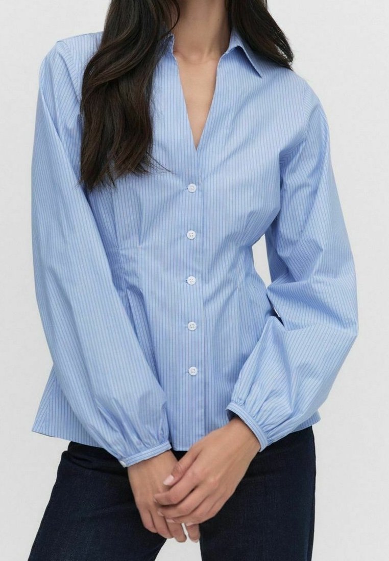 Femme portant une chemise ajustée bleu clair rayée boutonnée avec de longues manches bouffantes et un pantalon foncé, mains jointes devant.
