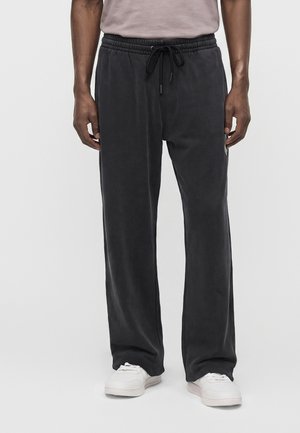 Homme portant un pantalon noir ample à cordon et des baskets blanches, debout devant un fond clair uni.
