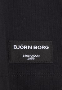 Černá látka s texturovaným povrchem, na němž je obdélníková nálepka s nápisy "BJÖRN BORG", "STOCKHOLM" a "1956" v bílé barvě.