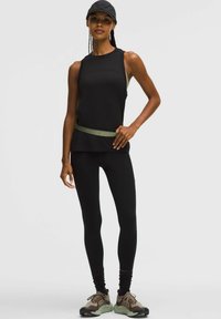 Vrouw in een zwart mouwloos shirt, zwarte leggings, een zwarte pet en bruin sportschoenen, staand met één hand op de heup tegen een effen achtergrond.