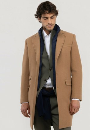 Hombre con abrigo marrón superpuesto, chaleco azul marino, chaqueta de traje oliva, camisa blanca y pantalones oscuros sobre fondo liso, mirando hacia abajo.