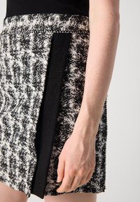 Juoda ir balta tweed sijonas su šonine išpjova ir lygia juoda detale. Medžiaga turi tekstūruotą paviršių, pabrėžiantį raštą.