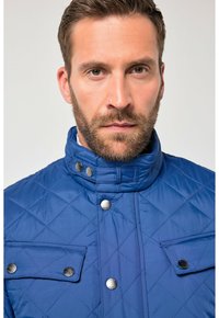 Niebieska pikowana kurtka z wysokim kołnierzem, srebrnymi zatrzaskami i dwoma przednimi kieszeniami. Charakteryzuje się wzorem w romby i gładką fakturą.