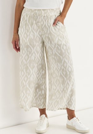 Kvinde iført cremefarvede bukser med vide ben med hvidt abstrakt mønster, kombineret med hvide sneakers og en hvid top, stående mod en hvid væg.