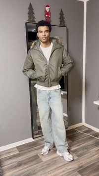 Casaco bomber em verde-oliva com capuz forrado a pelo, combinado com jeans azul-claro e ténis brancos. Em frente a um espelho, ambiente interior.