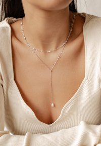 Naszyjnik warstwowy ze srebra, z delikatnymi łańcuszkami, małymi koralikami i pojedynczym zawieszką w kształcie perły. Noszony z kremowym, prążkowanym topem.