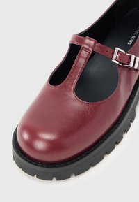 Μπορντό δερμάτινη παπούτσα Mary Jane με στρογγυλή μύτη, ρυθμιζόμενη πόρπη στο πάνω μέρος και χοντρή μαύρη καουτσούκ σόλα.