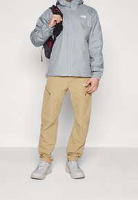 Vaaleanharmaa vedenpitävä takki hupulla, vetoketjullinen edestä ja logolla; beiget housut sivuvetoketjuilla; vaaleanharmaat kestävät kengät.