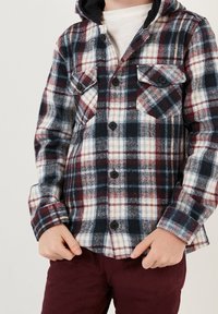 Chemise en flanelle à carreaux bleu marine, rouge et blanc avec deux poches poitrine et boutons noirs. Dispose d'une capuche et d'un motif texturé.