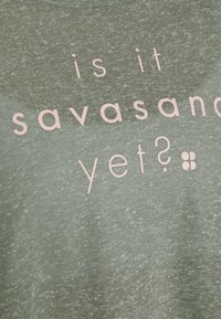 Olivově zelené bavlněné tričko s jemně růžovým textem "už je to savasana?" a malým květinovým detailem na konci.