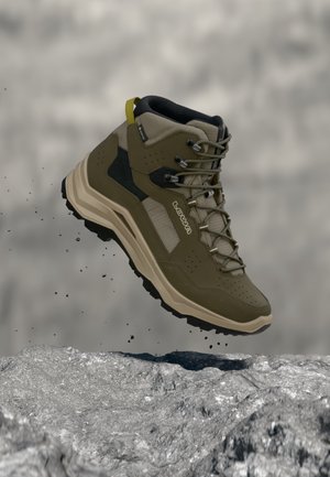 Olivově zelená turistická bota s černou podrážkou a tkaničkami, vznášející se nad kamenitým terénem s padajícími částicemi hlíny.