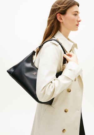 Jeune femme en trench-coat beige portant un sac à main en cuir noir avec des détails dorés sur l'épaule, tournée vers la droite.