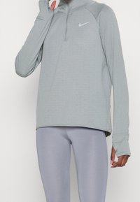 Lichtgrijze lange mouw sporttop met een halve rits, met een geweven patroon en duimopeningen. Gecombineerd met lichtgrijze leggings.