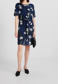 Mekko, jossa on kukkakuosisia tummansinisiä sävyjä, lyhyet hihat, joustava vyötärö ja rento istuvuus. Yhdistetty mustan clutch-laukun ja mustien mulojen kanssa.