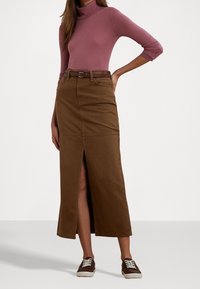 Kvinna iklädd en mauve polotröja med trekvartslång ärm, en brun midikjol med bälte och framficka, samt bruna sneakers med vita snören.